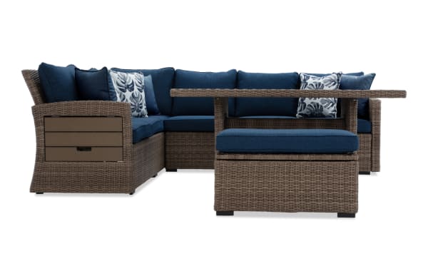 Outdoor sectional sofa set with brown wicker frame, navy blue cushions, and patterned throw pillows, featuring a matching ottoman and table.