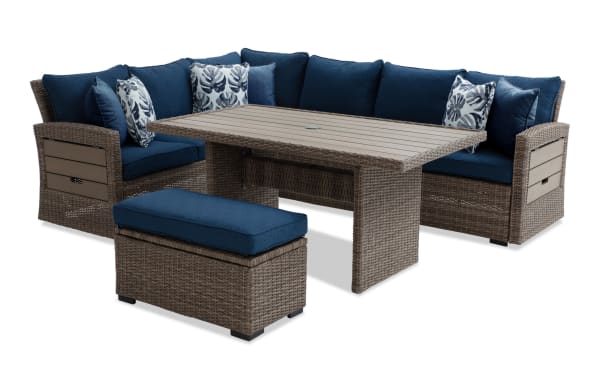 Outdoor patio sectional sofa set with brown wicker frame, navy blue cushions, floral throw pillows, and a matching wicker coffee table and ottoman.