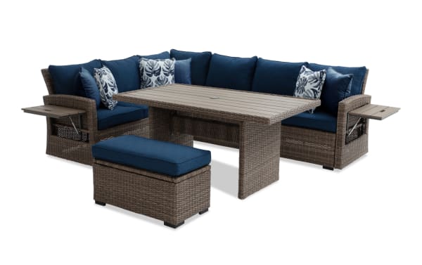Outdoor patio sectional sofa set with brown wicker frame, navy blue cushions, floral and solid throw pillows, rectangular wicker table, and matching ottoman.