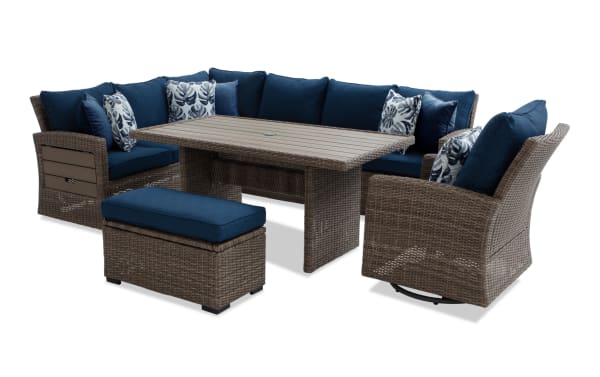 Outdoor patio furniture set featuring a brown wicker sectional sofa with navy blue cushions, a matching ottoman, swivel armchair, and a rectangular wicker dining table.