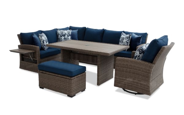 Outdoor patio furniture set with brown wicker frame, navy blue cushions, floral accent pillows, rectangular dining table, ottoman, and swivel chair.