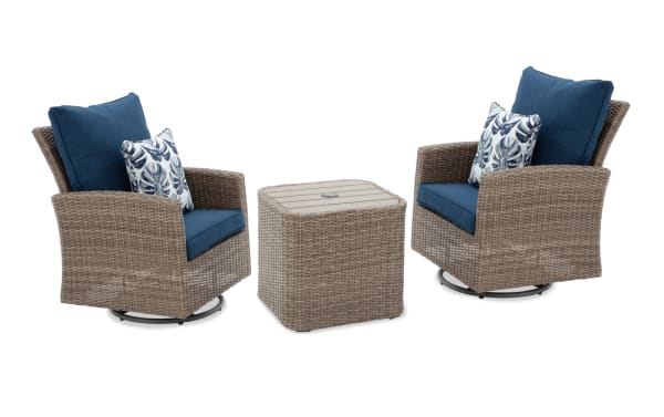 Outdoor patio set with two swivel wicker armchairs featuring blue cushions and tropical leaf patterned throw pillows, paired with a matching wicker side table with a slatted top.