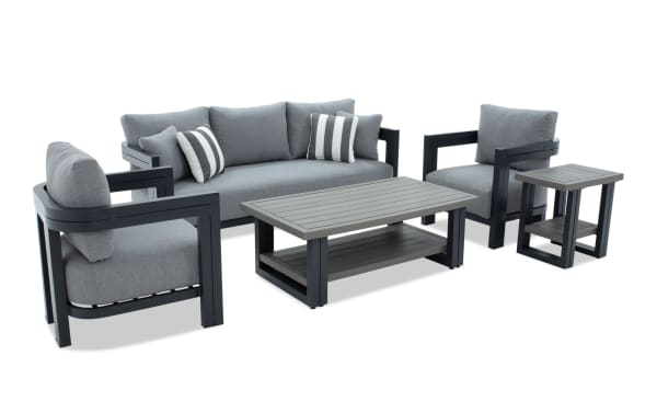 Modern outdoor patio furniture set with gray cushioned sofa, two armchairs, and matching coffee and side tables featuring black metal frames and slatted tabletops.
