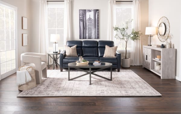 Modern living room with a dark blue leather sofa, glass-top metal coffee table, beige armchair, light wood sideboard, and large patterned area rug.