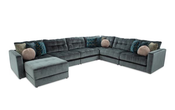 Large L-shaped sectional sofa in dark gray velvet with tufted cushions, includes matching ottoman and decorative throw pillows in various textures and colors.