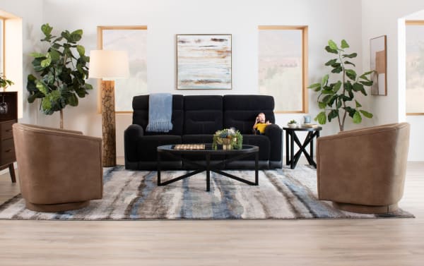 Modern living room with a black fabric sofa, two brown leather swivel chairs, a patterned area rug, round black coffee table, and indoor plants.