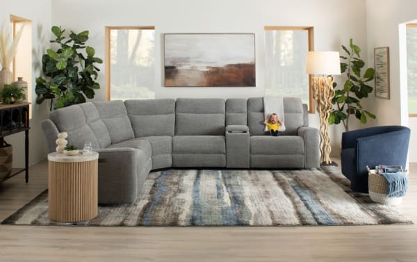 Modern living room with a gray fabric sectional sofa featuring reclining seats and a center console, complemented by a blue upholstered armchair, wooden side table, abstract wall art, and decorative plants.