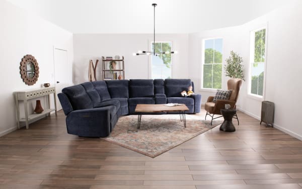 Modern living room with a navy blue fabric sectional sofa, wooden coffee table with metal legs, patterned area rug, tan leather armchair, and large windows with natural light.