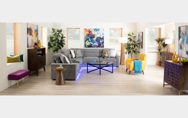 Modern living room with a large gray sectional sofa featuring blue LED lighting underneath, a yellow armchair with a blue throw, wooden side tables, colorful abstract wall art, and natural light from multiple windows.