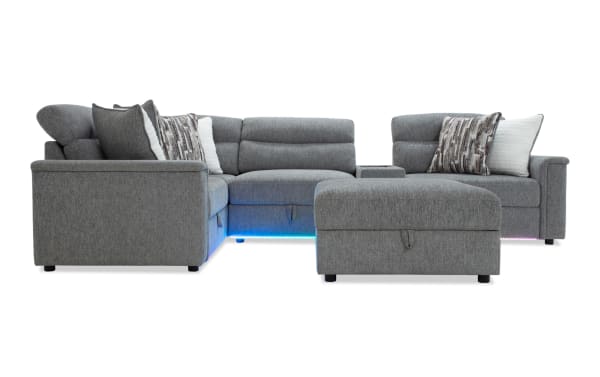 Modern gray fabric sectional sofa with chaise and ottoman, featuring plush cushions, patterned throw pillows, and built-in LED lighting underneath.