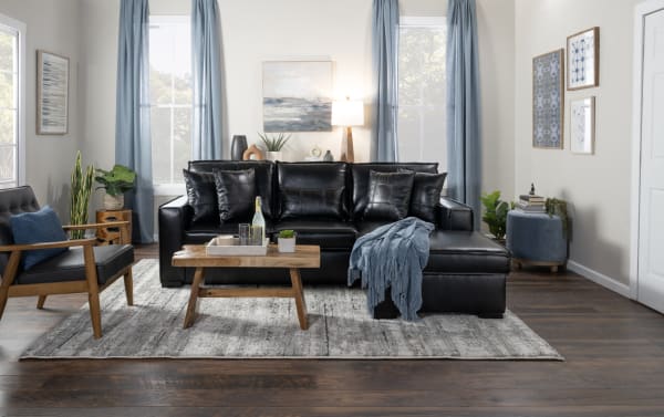 Modern living room with black leather sectional sofa, wooden coffee table, gray area rug, blue-gray curtains, and decorative plants.