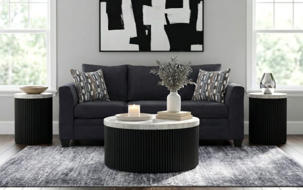 Modern living room setup with a dark gray fabric sofa, black ribbed round marble-top coffee and side tables, patterned throw pillows, and abstract black-and-white wall art.