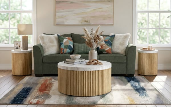 Living room setup with green fabric sofa, decorative patterned and fluffy pillows, round wooden coffee table and side tables with marble tops, neutral-toned rug with abstract patterns, and natural dried floral centerpiece.