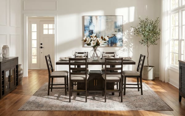 Dining room with dark wood table and six cushioned chairs on a patterned area rug, featuring a glass vase with white flowers and abstract blue wall art.