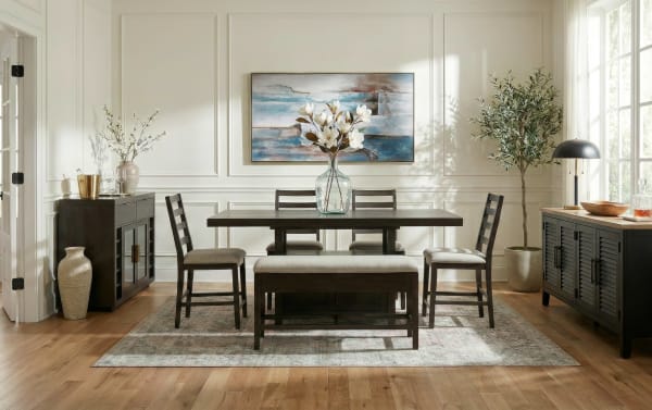 Modern dining room set with dark wood rectangular table, cushioned bench, and four ladder-back chairs on patterned rug, bright natural light.