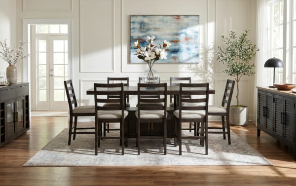 Dark wood dining table set with eight cushioned ladder-back chairs on a patterned area rug, accented by floral centerpiece, wall art, and natural light.