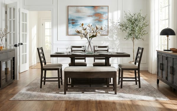 Modern dining room with dark wood table, four matching ladder-back chairs, and a cushioned bench on a patterned rug, accented by a floral centerpiece, abstract wall art, and natural light from large windows.