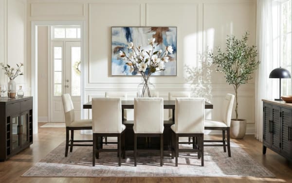 Modern dining room with dark wood rectangular table, eight beige upholstered chairs, floral centerpiece, abstract wall art, and natural light.
