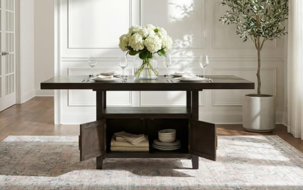 Dark wood rectangular dining table with open storage cabinet, set with white plates, glassware, beige napkins, and a vase of white hydrangeas on a patterned rug in a bright room.