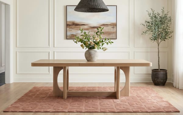 Modern rectangular wooden dining table with rounded edges on a textured terracotta rug, featuring a ceramic vase with green foliage and flowers, set in a bright room with white paneled walls, a framed landscape painting, a black woven pendant light, and a potted olive tree.