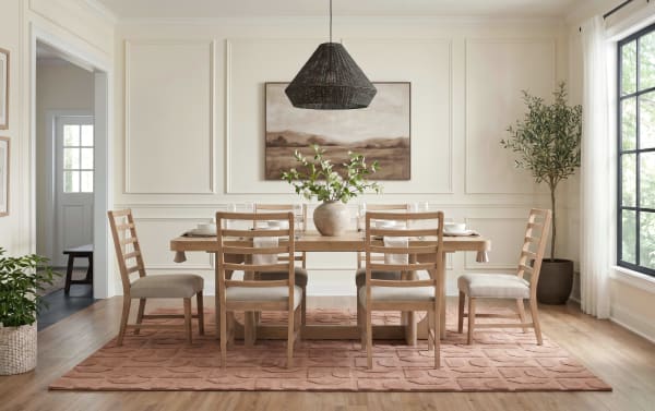 Dining room with a natural wood table and six matching ladder-back chairs with beige cushions, a textured terracotta rug, a ceramic vase with green foliage centerpiece, a large window with white curtains, and a black woven pendant light.