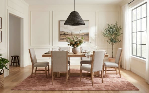 Modern dining room with a wooden table and six beige upholstered chairs on a textured pink area rug, featuring a large window, potted plants, and a woven black pendant light.