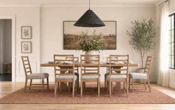 Dining room with a wooden rectangular table and six matching ladder-back chairs with beige cushions, a large terracotta vase with greenery centerpiece, a woven black pendant light, a textured rust-colored rug, botanical wall art, and a tall potted olive tree near a window with sheer curtains.