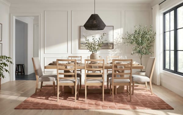Modern dining room with a light wood rectangular table, eight beige cushioned wooden chairs, a textured rust-colored rug, large window, and decorative plants.