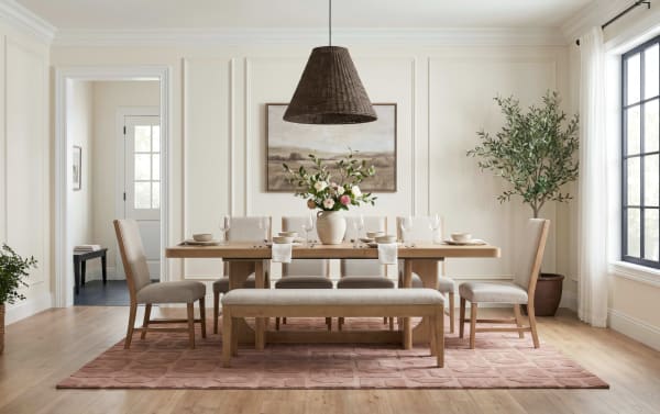 Modern dining room with light wood table, beige upholstered chairs and bench, woven pendant light, floral centerpiece, and large window.