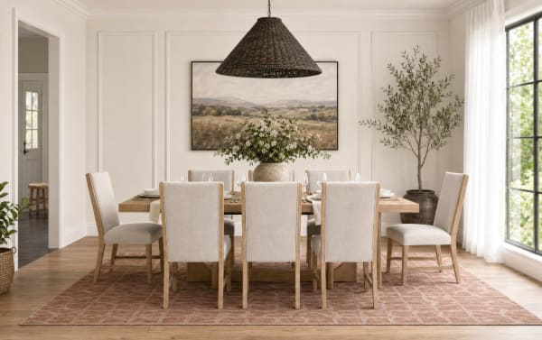Dining room with a wooden table and eight beige upholstered chairs on a patterned rust-colored rug, featuring a large woven pendant light, a vase with white flowers, a landscape painting, and natural light from a large window.