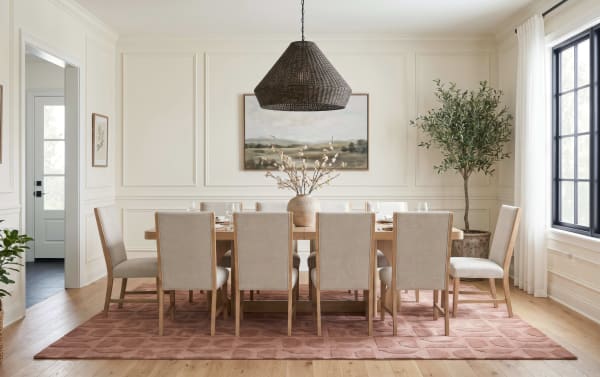 Modern dining room with a large wooden table, ten beige upholstered chairs, a textured rust-colored area rug, a woven pendant light, and neutral wall art.
