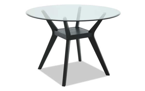 Round dining table with a clear glass top and black wooden tripod legs, featuring a modern minimalist design.