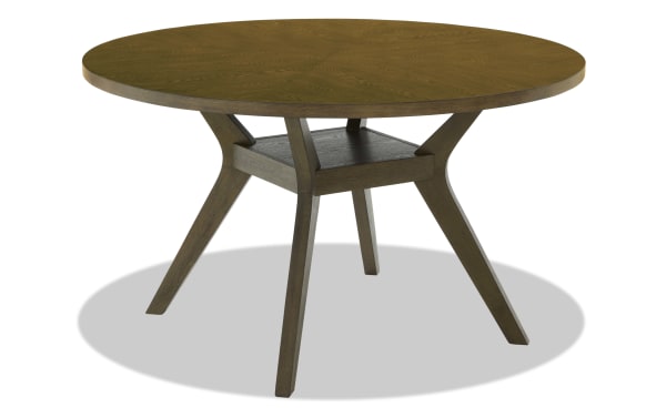 Round wooden dining table with a medium brown finish, featuring angled legs and a small central shelf for additional storage or decor.