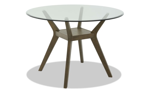 Round dining table with clear glass top and dark wood angled legs, featuring a small wooden shelf underneath for storage or decor.