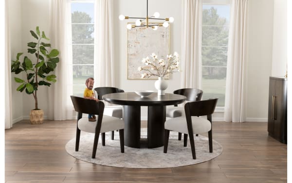 Modern dining room with round dark wood table, four cushioned chairs with dark wooden frames, beige area rug, large windows with white curtains, decorative white vase with flowers, and a potted green plant on wooden floor.