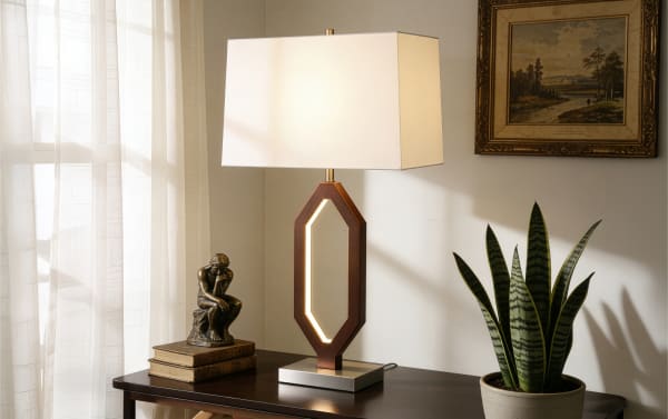 Modern table lamp with a rectangular white fabric shade and geometric wooden base with built-in LED light, placed on a dark wood surface next to a potted snake plant and bronze sculpture.