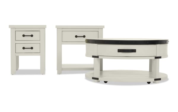Set of three white wooden tables with black metal handles and accents, including a two-drawer nightstand, a single-drawer side table, and a round coffee table with a lower shelf.