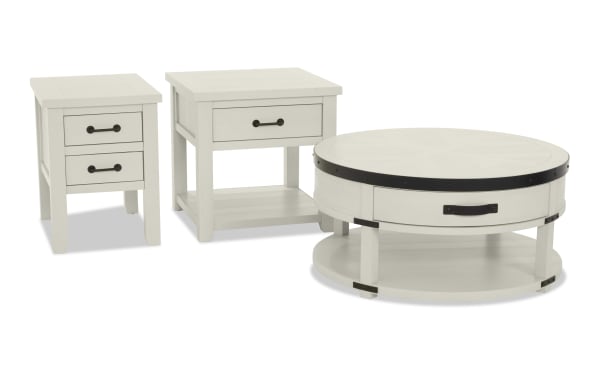 Set of three white wooden tables including a small two-drawer side table, a larger single-drawer end table, and a round coffee table with black metal accents and lower shelf.