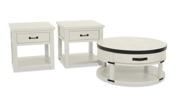 Set of three white wooden tables including two square end tables with single drawers and a round coffee table with black metal accents and handles, featuring lower shelves for storage.