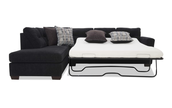 Black fabric sectional sofa with a pull-out metal frame sleeper bed, white mattress, and four decorative pillows in patterned and solid colors.