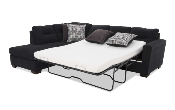 Black fabric sectional sofa with a pull-out metal frame bed and white mattress, featuring patterned and solid throw pillows for added comfort.