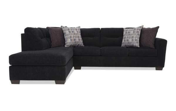 Black fabric sectional sofa with tufted back cushions, chaise lounge on left side, and four decorative pillows in patterned and solid dark colors.