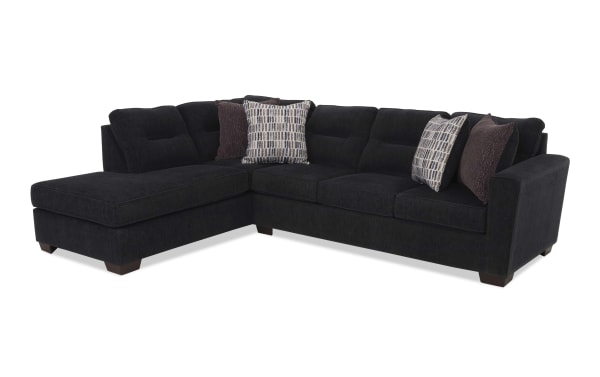 Black fabric sectional sofa with chaise lounge, tufted back cushions, wooden legs, and decorative patterned and solid throw pillows.