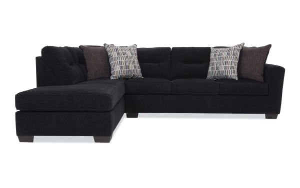 Modern black fabric sectional sofa with tufted back cushions, L-shaped design, wooden legs, and four decorative throw pillows in patterned and solid brown fabric.