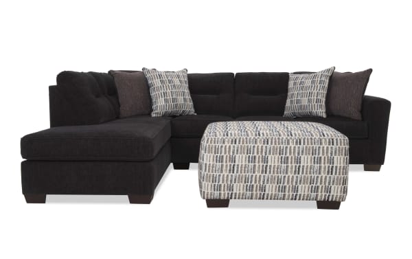 Dark gray fabric sectional sofa with tufted back cushions, patterned beige and gray ottoman, and matching decorative pillows, modern style.