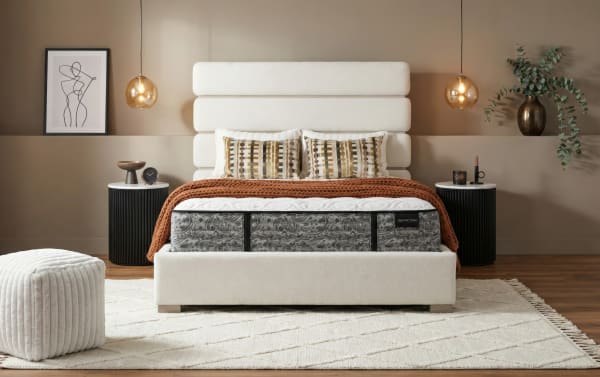Modern bedroom featuring a Distinction mattress with a white upholstered bed frame, textured brown throw, patterned pillows, black round nightstands, and warm pendant lighting.