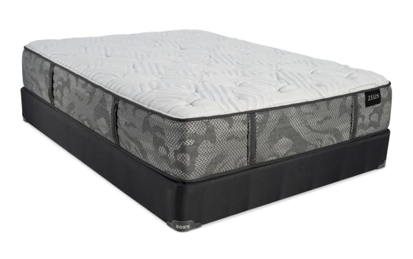 Queen-size Zeus mattress with a plush white quilted top and gray patterned sides, resting on a black fabric foundation labeled "Bob's."