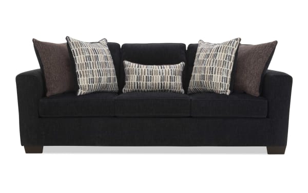 Black fabric sofa with three seat cushions and five decorative pillows in brown and patterned beige tones, featuring a modern, boxy design and wooden legs.