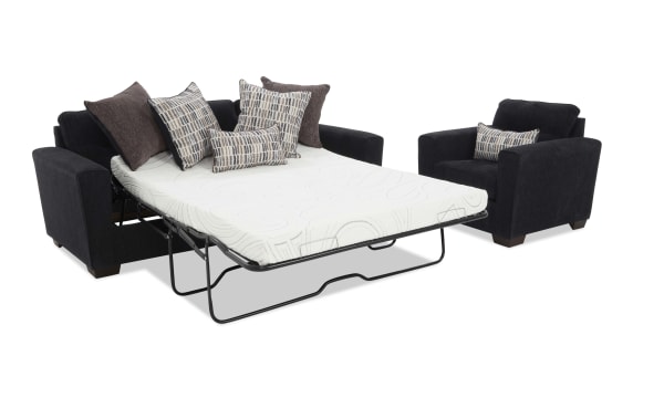 Black fabric sleeper sofa with a white mattress extended, featuring multiple patterned and solid brown cushions, paired with a matching black armchair with a patterned cushion.