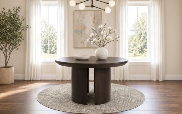 Round dark wood dining table with a textured beige circular rug, white vase with magnolia flowers, neutral curtains, and natural light from windows.